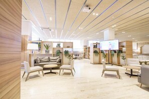 Un moderno vestíbulo de hotel con una zona de estar central, una pantalla de televisión e iluminación lineal en el techo.
