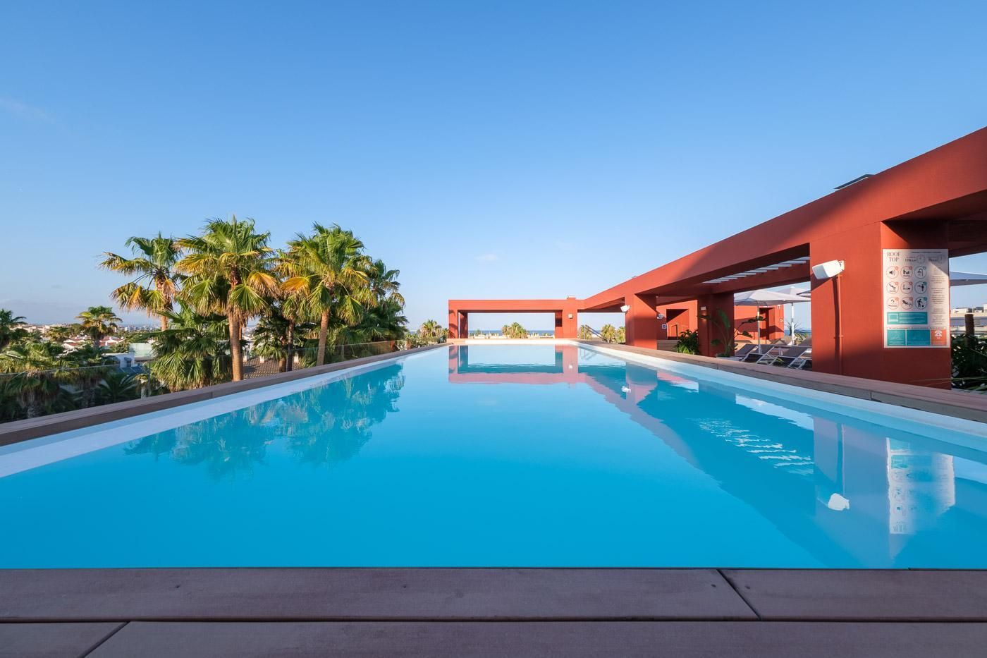 Una piscina azul cristalina con una estructura roja moderna, palmeras y un cielo despejado.
