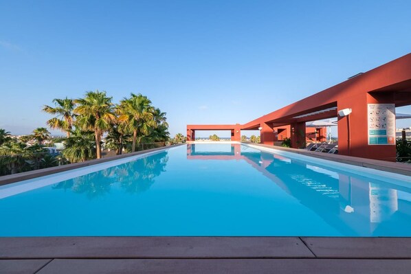 Una piscina azul cristalina con una estructura roja moderna, palmeras y un cielo despejado.