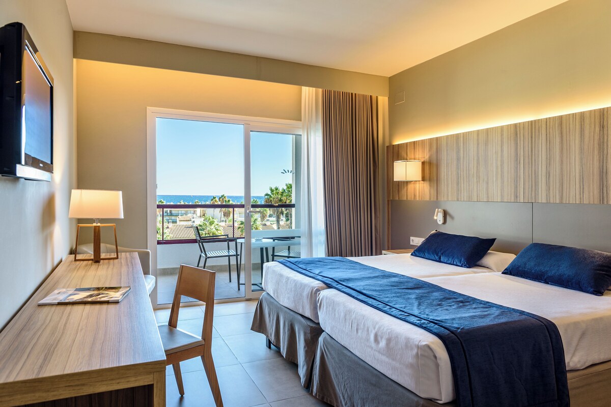Habitación de hotel con cama, escritorio, silla, televisor, balcón con vistas y lámpara.