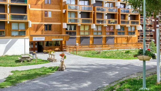 A multi-story wooden building with balconies, a covered entrance, and a gravel pathway leading to a picnic area.