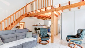 Amplia sala de estar con un sofá gris, dos mecedoras, una escalera de madera y una cocina al fondo.