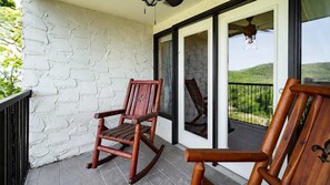 Une chaise berçante en bois sur un balcon, avec un mur en briques blanches et une porte vitrée donnant sur un paysage verdoyant.