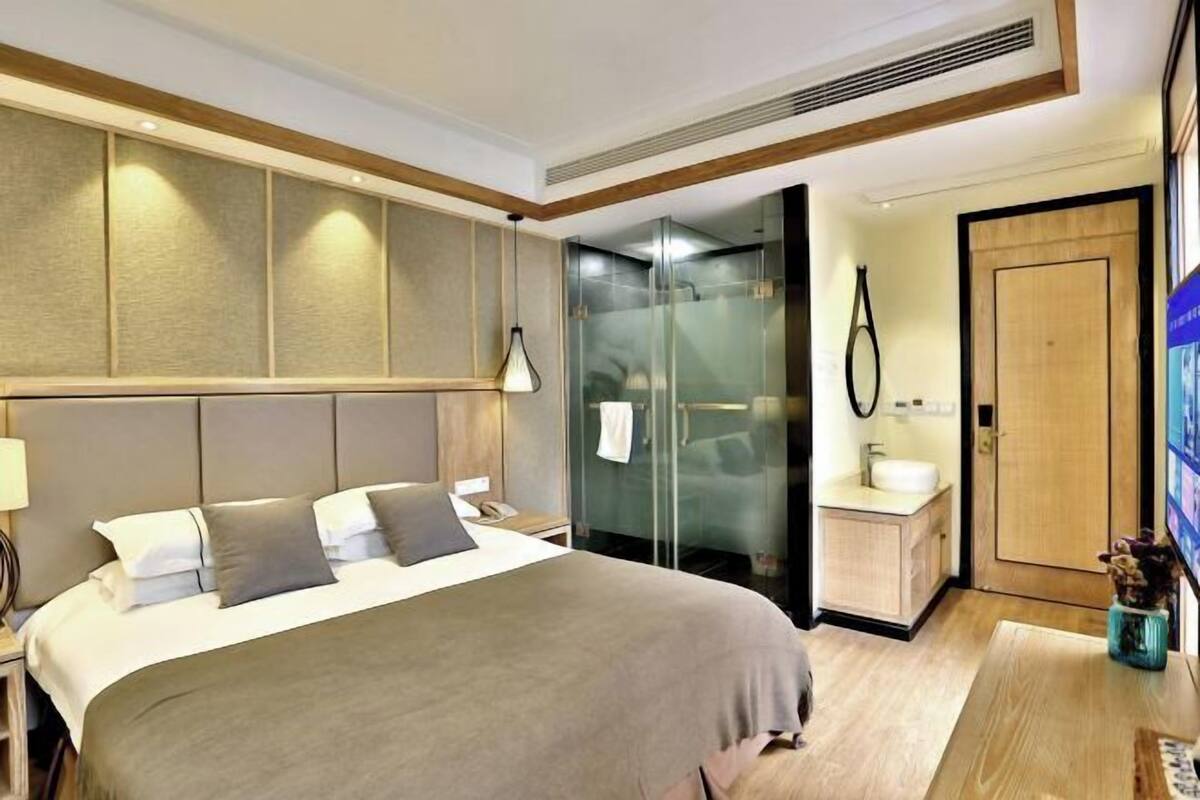 Une chambre d’hôtel comprenant un lit, une table de chevet, une commode, une cabine de douche en verre, un meuble en bois et une petite télévision.