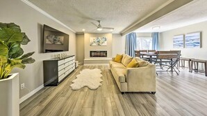 A modern living room with a sofa, a coffee table, and a fireplace. There is a dining area with a table and chairs. - The Harold House  (Olathe)