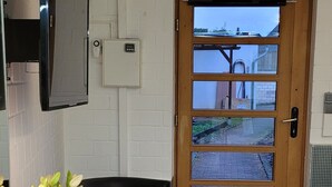 Een compacte kamer met een houten deur, een zwarte bank, een witte stoel, een klein tafeltje met een plant en een spiegel aan de muur.