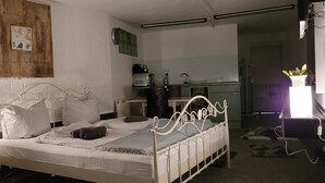 Een slaapkamer met een bed met een wit metalen frame, een nachtkastje, een wandplank met keukenspullen.