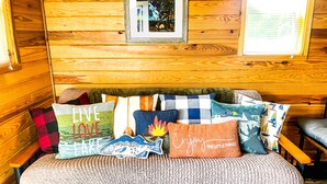 Ein Holzblockhaus-Interieur mit einer Couch, geschmückt mit verschiedenen Dekokissen, einem gerahmten Bild an der Wand und einem Fenster mit Jalousien.