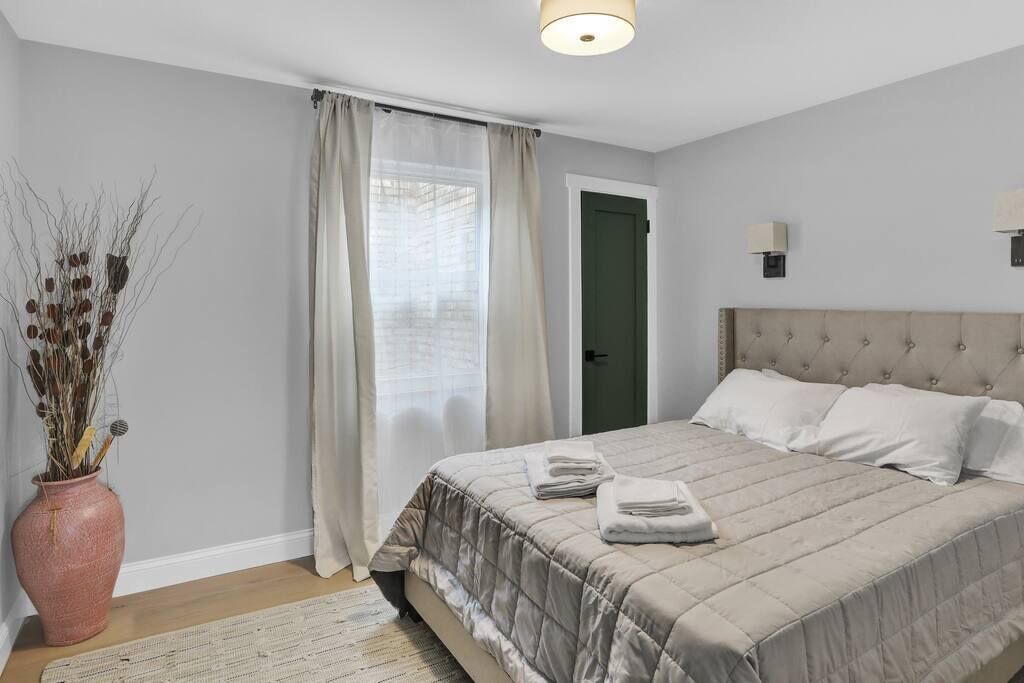 Une chambre à coucher avec un lit, des rideaux, un vase contenant des plantes séchées et une porte.