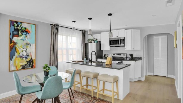 A modern kitchen with white cabinets, a central island with stools, a dining area with a glass table and chairs, and a large abstract painting on the wall. - Luxury Apartment In Lakewood (Lakewood)