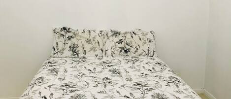 A bedroom with a bed featuring a floral-patterned bedspread, a framed landscape painting on the wall, and a plain white wall.