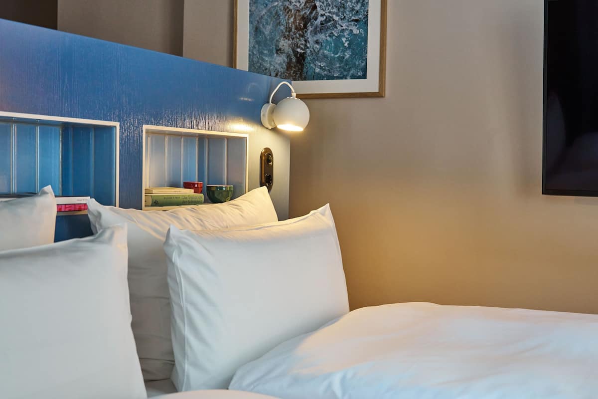 A hotel room with a bed, pillows, a nightstand with books, a lamp, and a painting on the wall.
