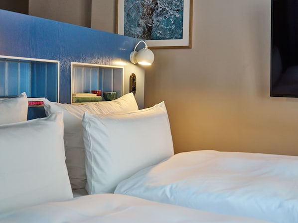 Habitación de hotel con cama, almohadas, mesita de noche con libros, lámpara y un cuadro en la pared.