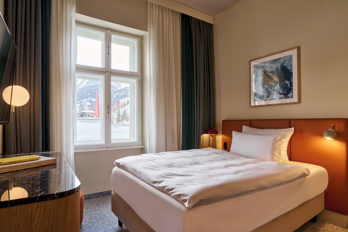A hotel room with a large bed, a desk with a lamp, a painting on the wall, and a window with curtains.