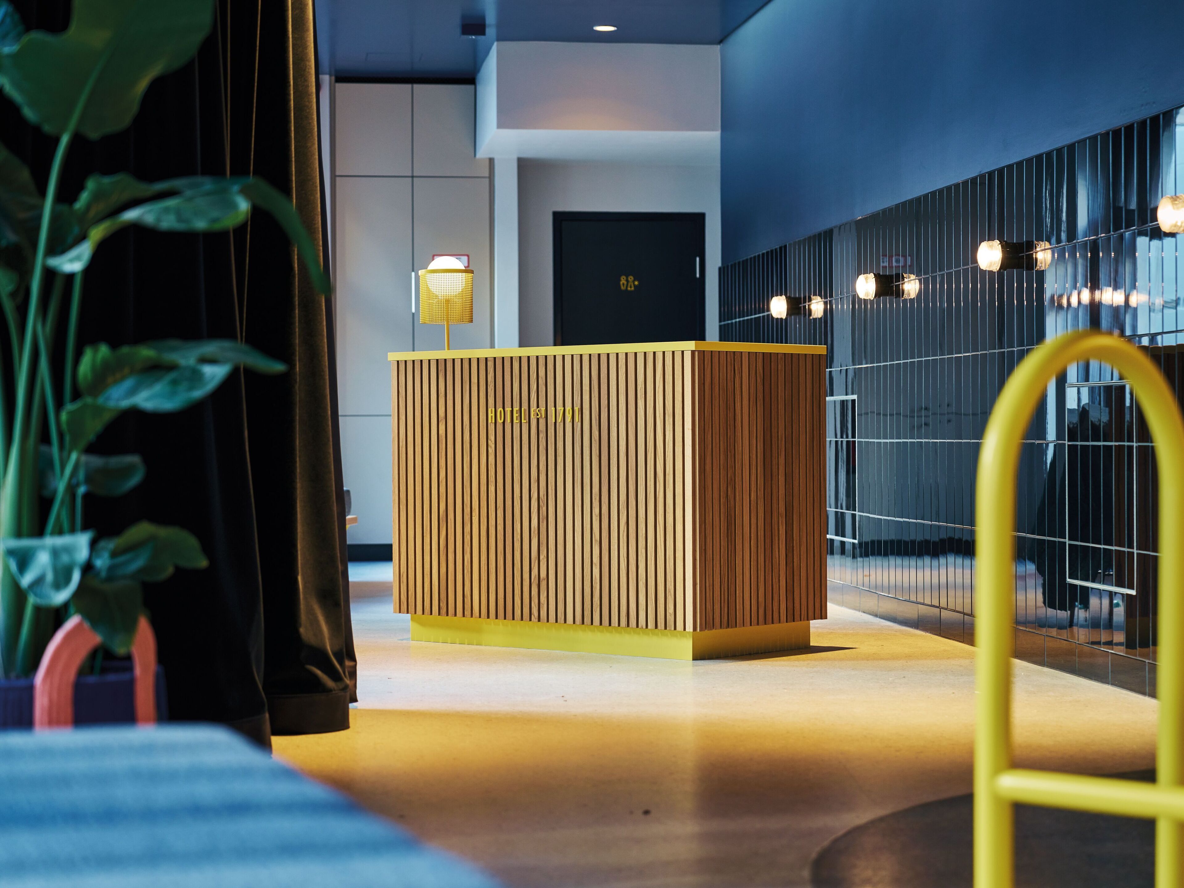 Un comptoir d’accueil avec des lambris en bois verticaux et un plateau jaune, un luminaire moderne et une rambarde en métal au premier plan.