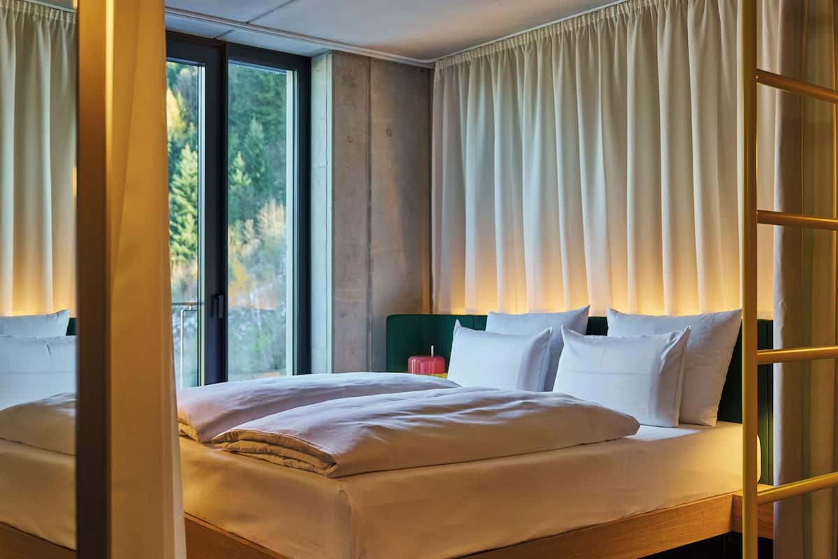 A modern hotel room with a large bed, a green headboard, and a view of trees through the window.