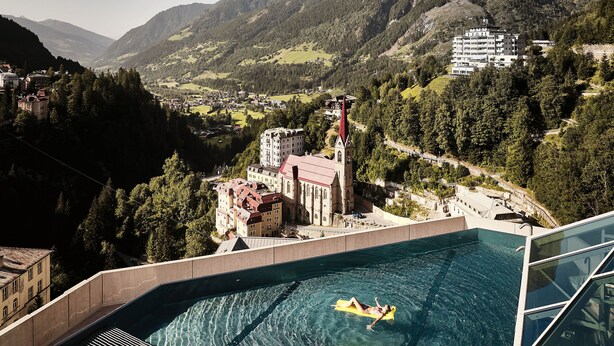 Una piscina con una persona en un flotador, rodeada de montañas y una iglesia.
