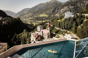 Una piscina con una persona en un flotador, rodeada de montañas y una iglesia.