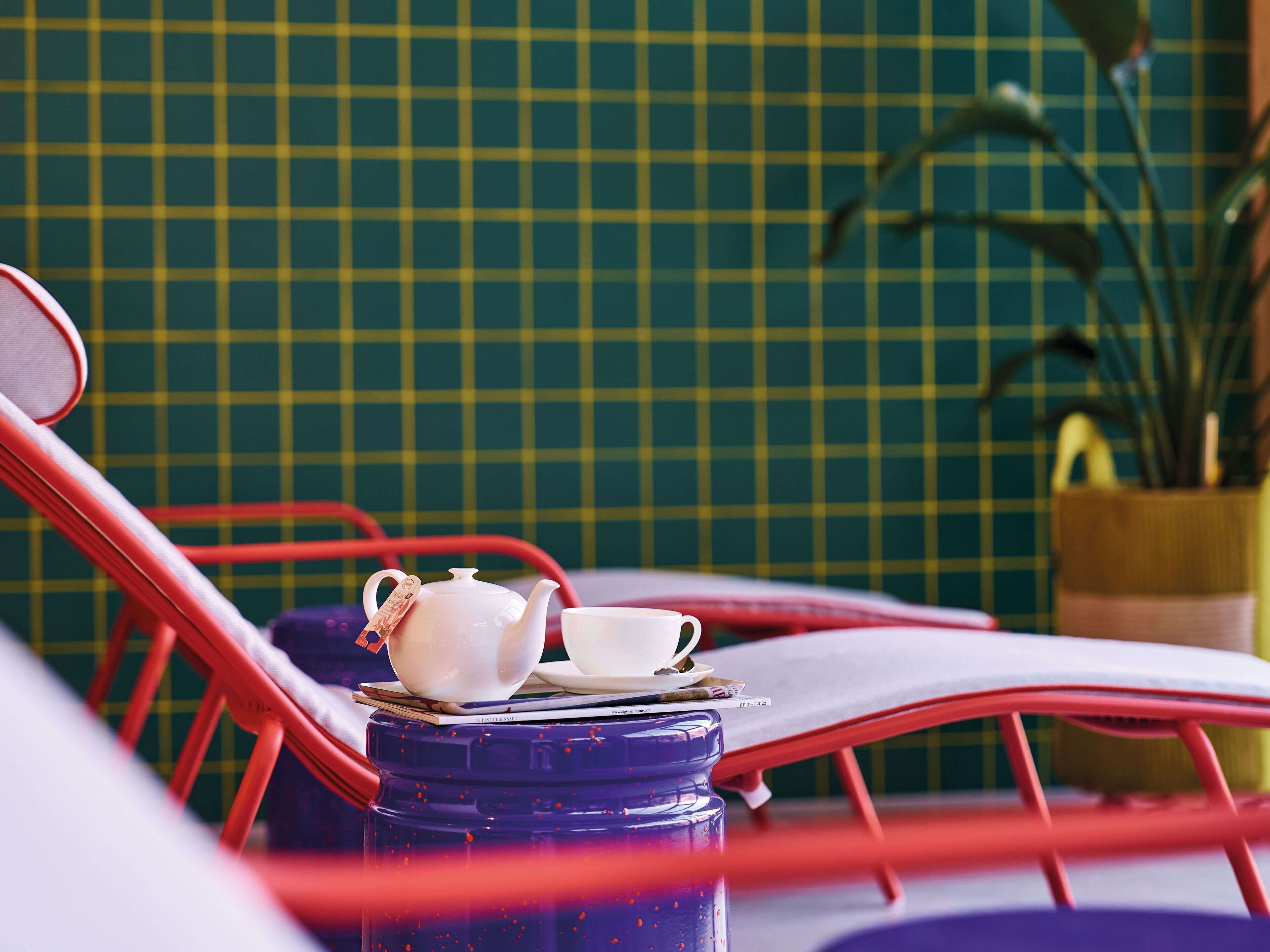 A red metal chair with a white teapot and cup on a table, a blue water bottle, and a green tiled wall in the background.