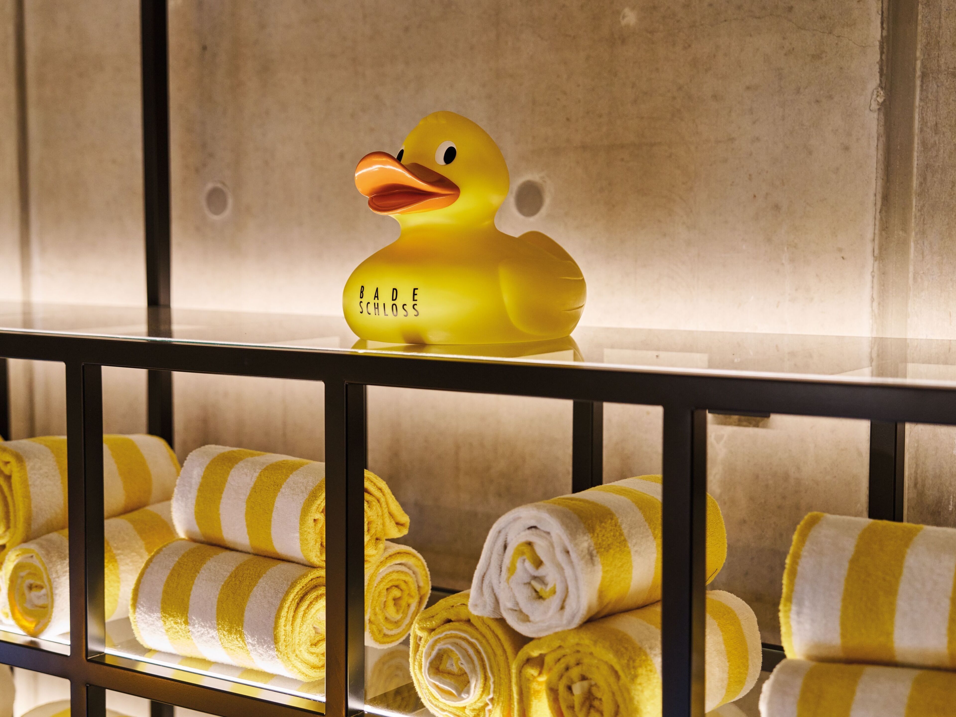 A yellow duck-shaped bath mat with "BADE SCHLOSS" text, and neatly rolled towels in yellow and white stripes.