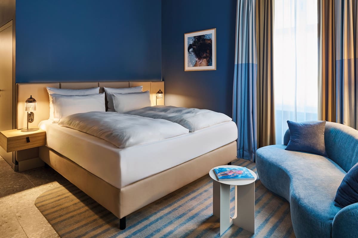 A neatly arranged hotel room with a large bed, a side table with a lamp, a small round table with a book, a blue armchair, and a window with curtains.