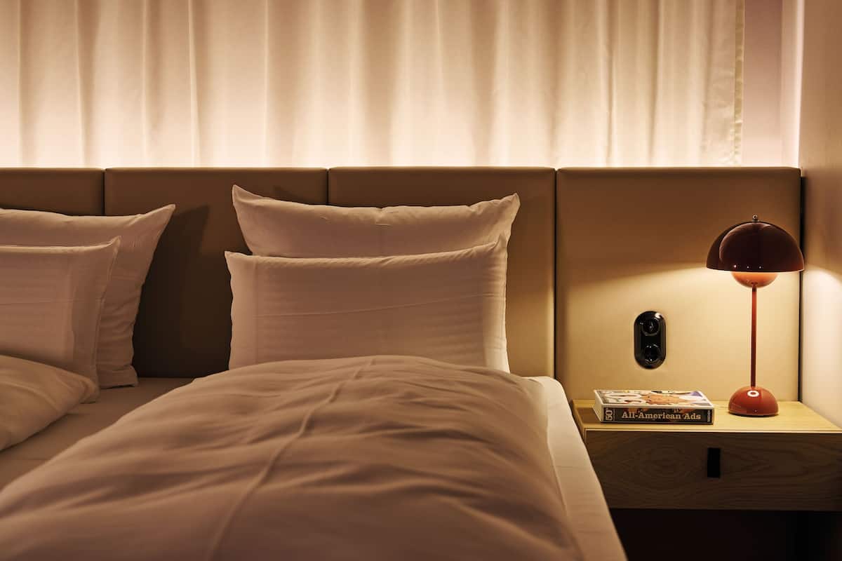 A hotel room with a bed, pillows, a nightstand with a lamp, and a book titled "AI-American Ads".