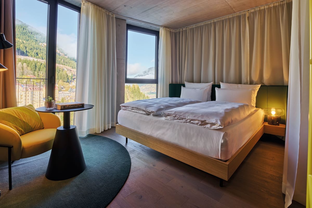 A hotel room with a large bed, a small table with a lamp, a sofa, and a view of a mountainous landscape.