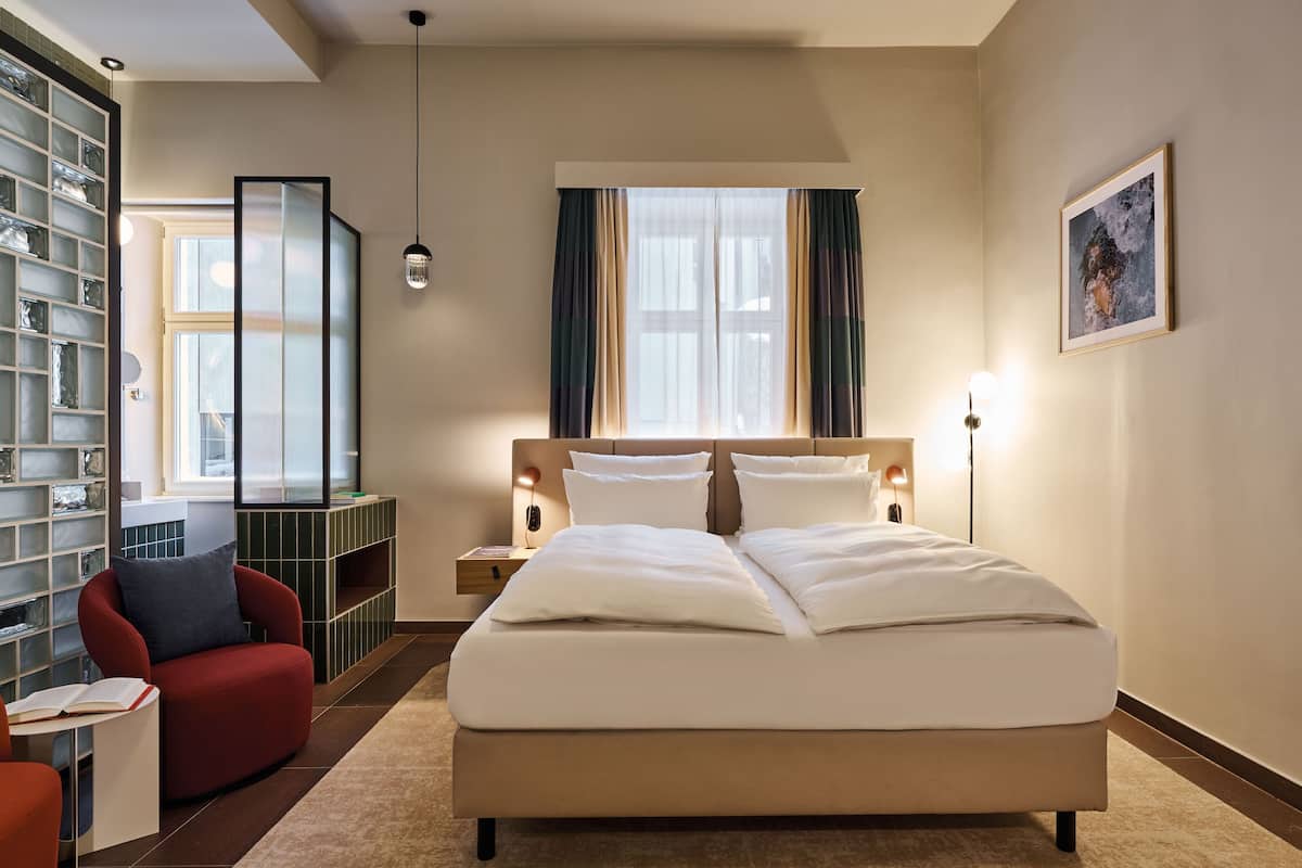 A hotel room with a large bed, a red armchair, a small table with a book, a bookshelf, a window with curtains, and a painting on the wall.