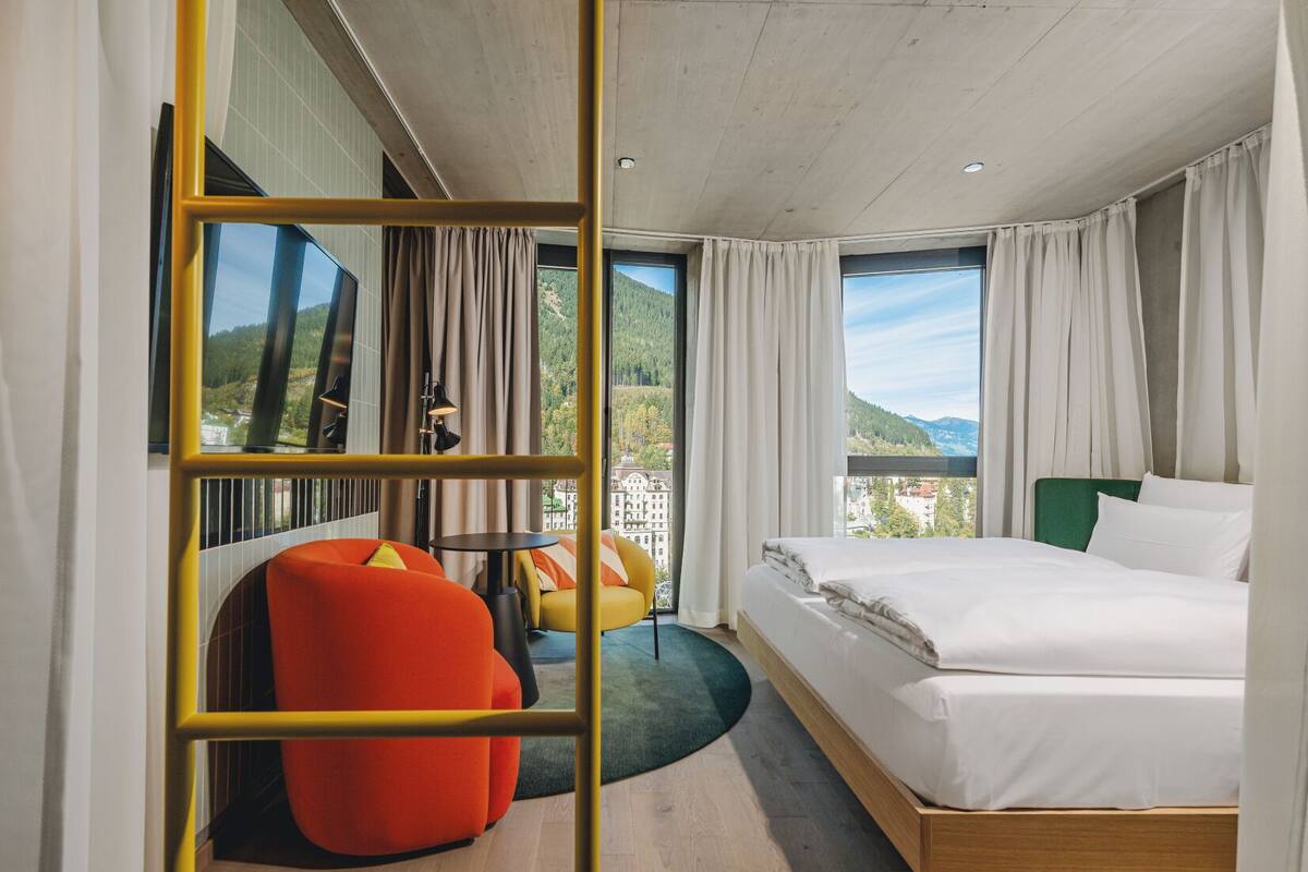 A hotel room with a bed, a red chair, a small table, and a view of a mountain.
