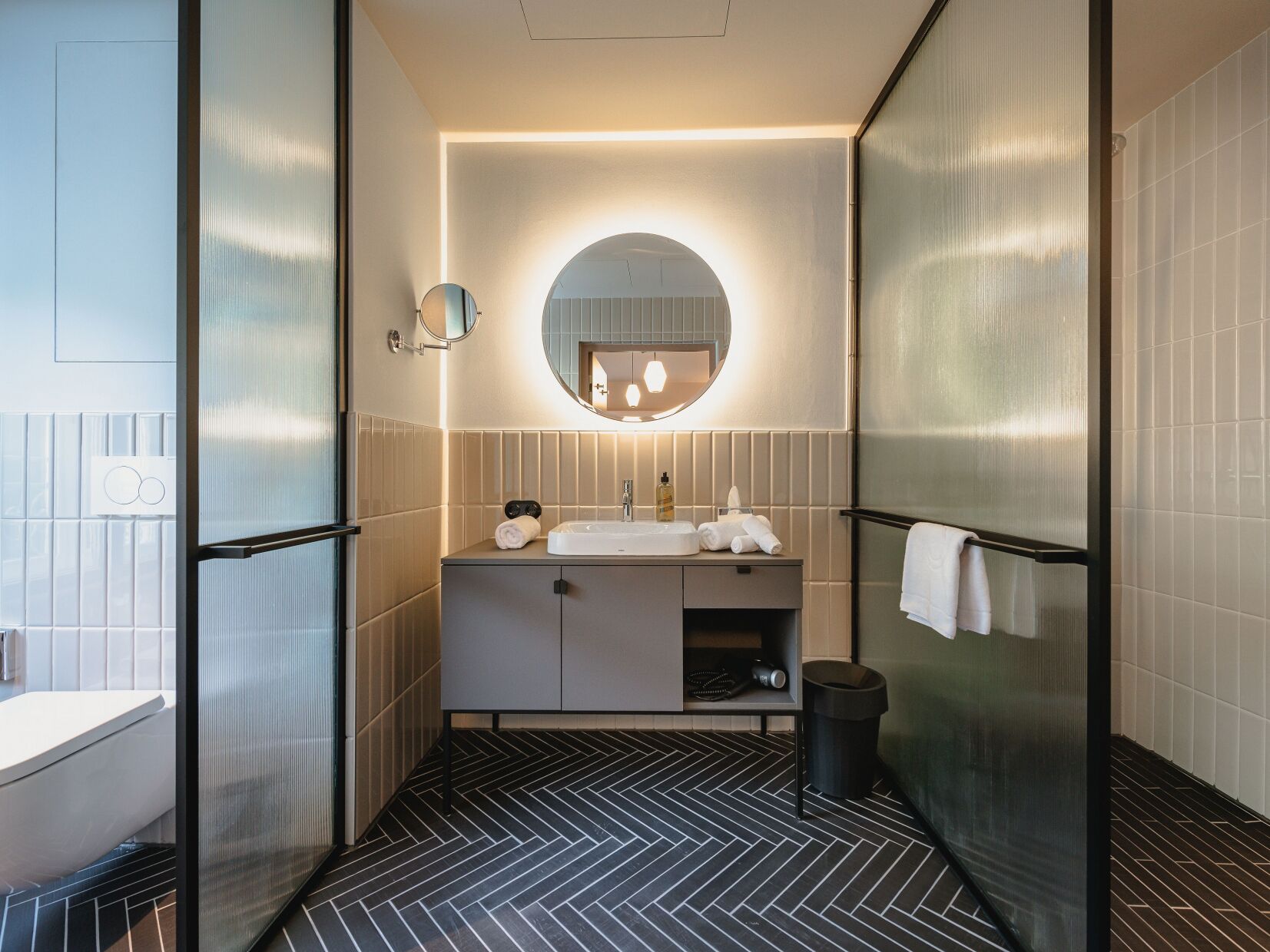 Un baño moderno con suelo de espiga en blanco y negro, un espejo redondo sobre el lavabo y un perchero en la pared.
