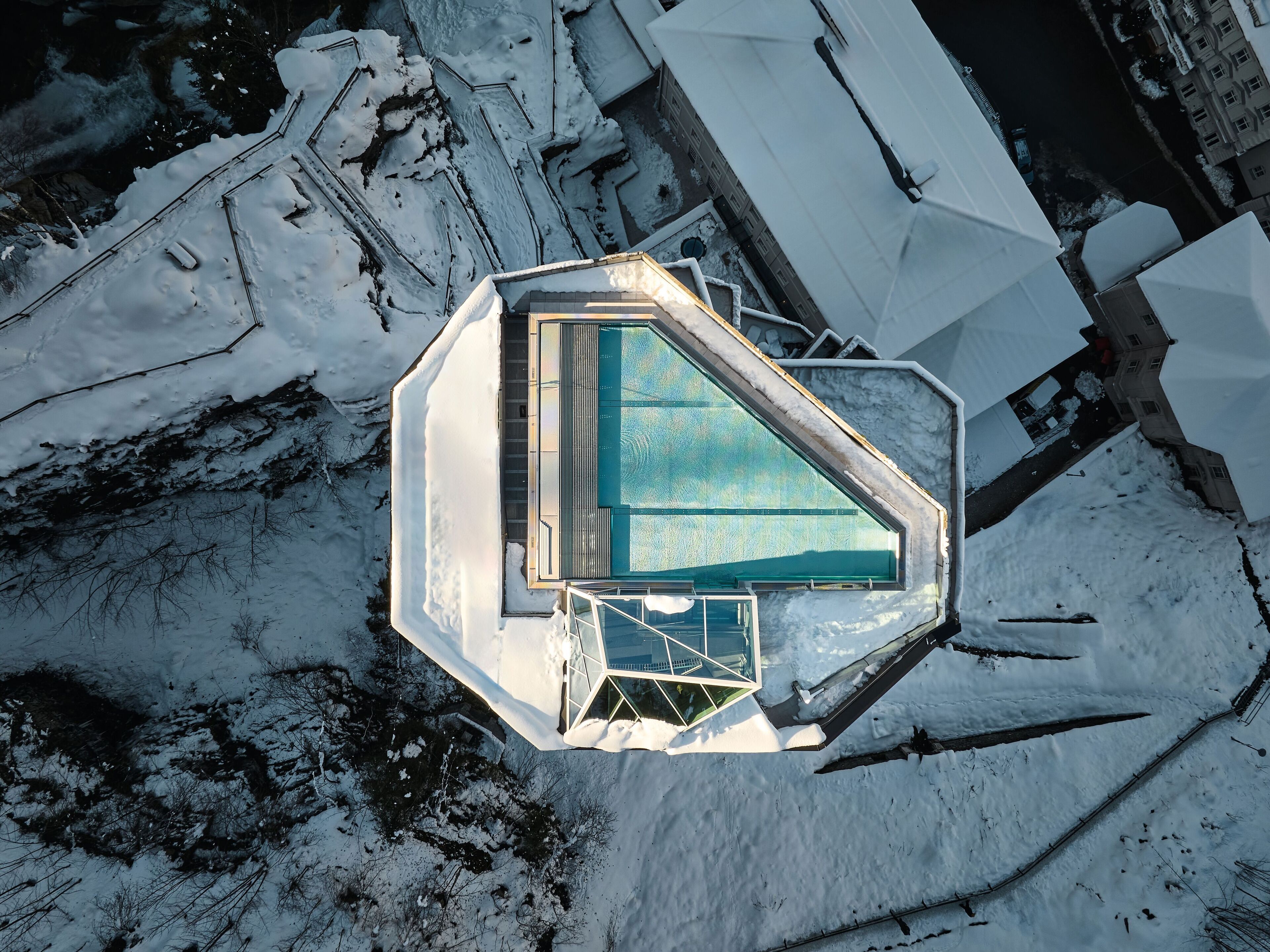 An aerial view of a modern building with a glass-enclosed swimming pool, surrounded by snow.