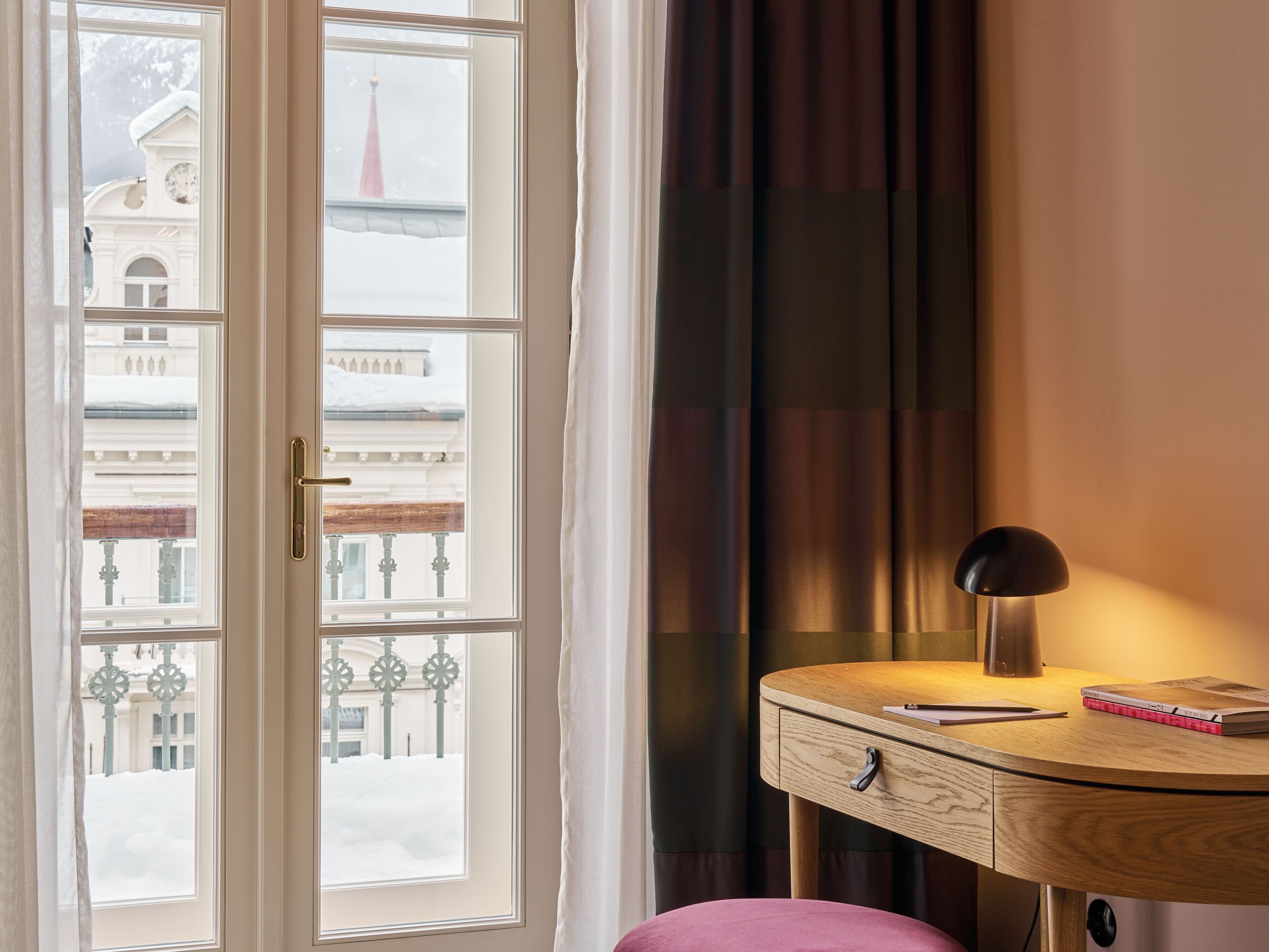 Una habitación con un ventanal grande, un escritorio con lámpara y una silla rosa.