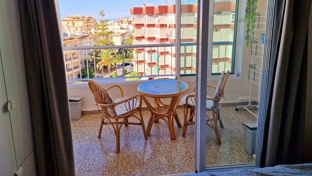 A balcony with a round table and wicker chairs, offering a view of a residential area with buildings and a clear sky. - Cozy apartment with pool (Nerja)