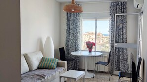 A compact living space with a sofa, a small table, a chair, a potted plant, and a view of buildings through large windows. - Cozy apartment with pool (Nerja)
