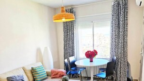A living room with a sofa, coffee table, and a dining table with chairs. There is a TV stand with a TV and a remote control. The room has a window with patterned curtains and a hanging light fixture. - Cozy apartment with pool (Nerja)