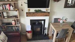 A cozy living room with a fireplace, a TV mounted above it, a dining table with chairs, and a bookshelf filled with books and decorative items.