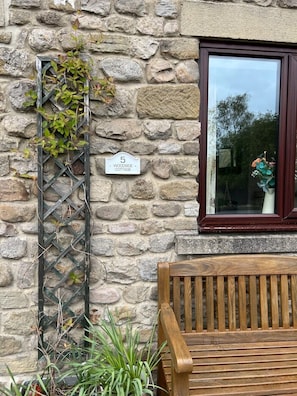 Un muro de piedra con un banco de madera y una celosía metálica con plantas trepadoras. Se ve una ventana con marco oscuro y un letrero que dice "5 WOODSIDE COTTAGE".