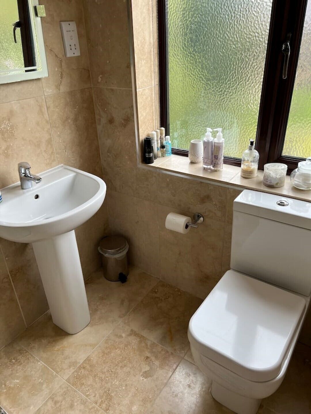 Ein Badezimmer mit einem Standwaschbecken, Toilette und einem Fenster mit Pflegeprodukten auf der Fensterbank.