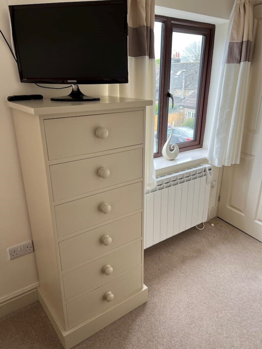 A room with a white dresser, a television on top, a window with curtains, and a radiator.
