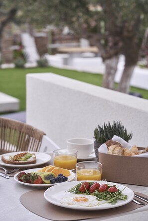 Un desayuno con platos de comida, un frutero y un vaso de jugo en una mesa al aire libre.