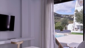 Habitación de hotel moderna con una televisión grande, una cama, una mesita y vistas a una piscina y casas en la colina.
