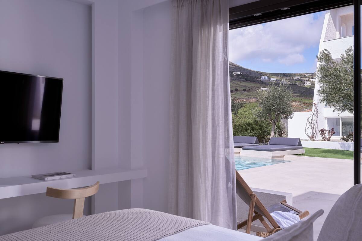 Quarto de hotel moderno com uma televisão grande, uma cama, uma mesa pequena e vista para uma piscina e casas na colina.