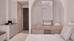 Un dormitorio moderno con una cama, un tocador con espejo, un lavabo y una pequeña mesa con una taza y un florero.