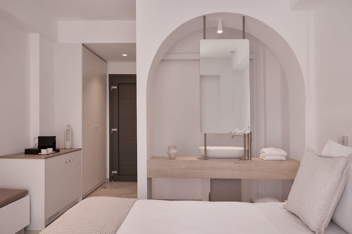 Um quarto moderno com uma cama, uma penteadeira com espelho, pia e uma pequena mesa com uma xícara e um vaso.