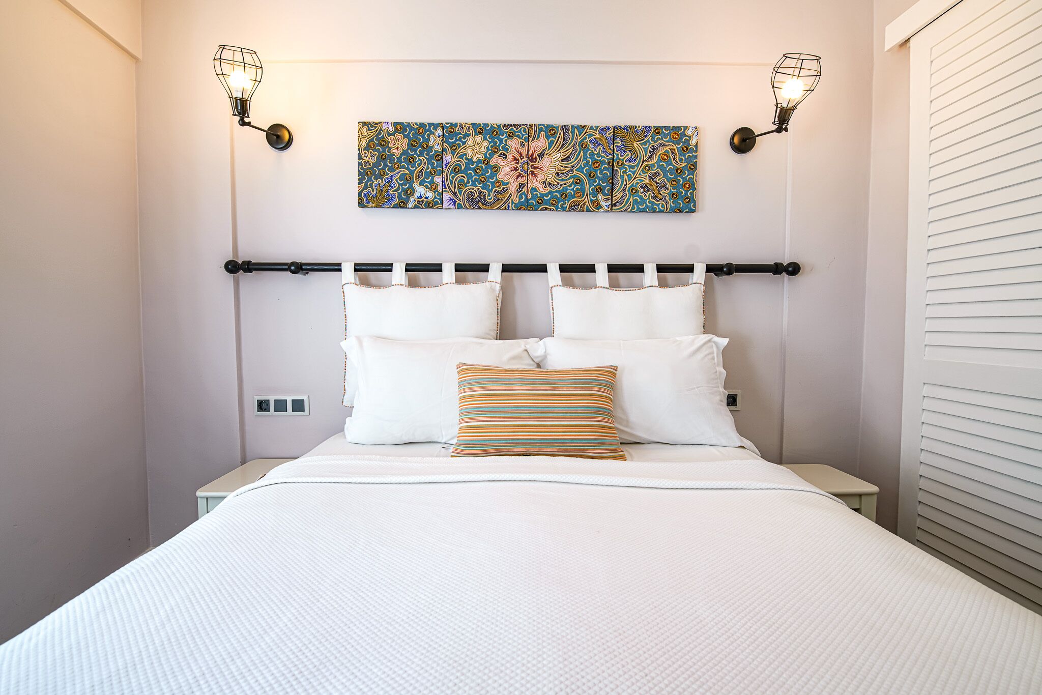 Ein ordentlich bezogenes Bett mit weißer Bettwäsche, einem farbenfrohen Streifenmuster-Kissen und einem Blumenbild darüber.