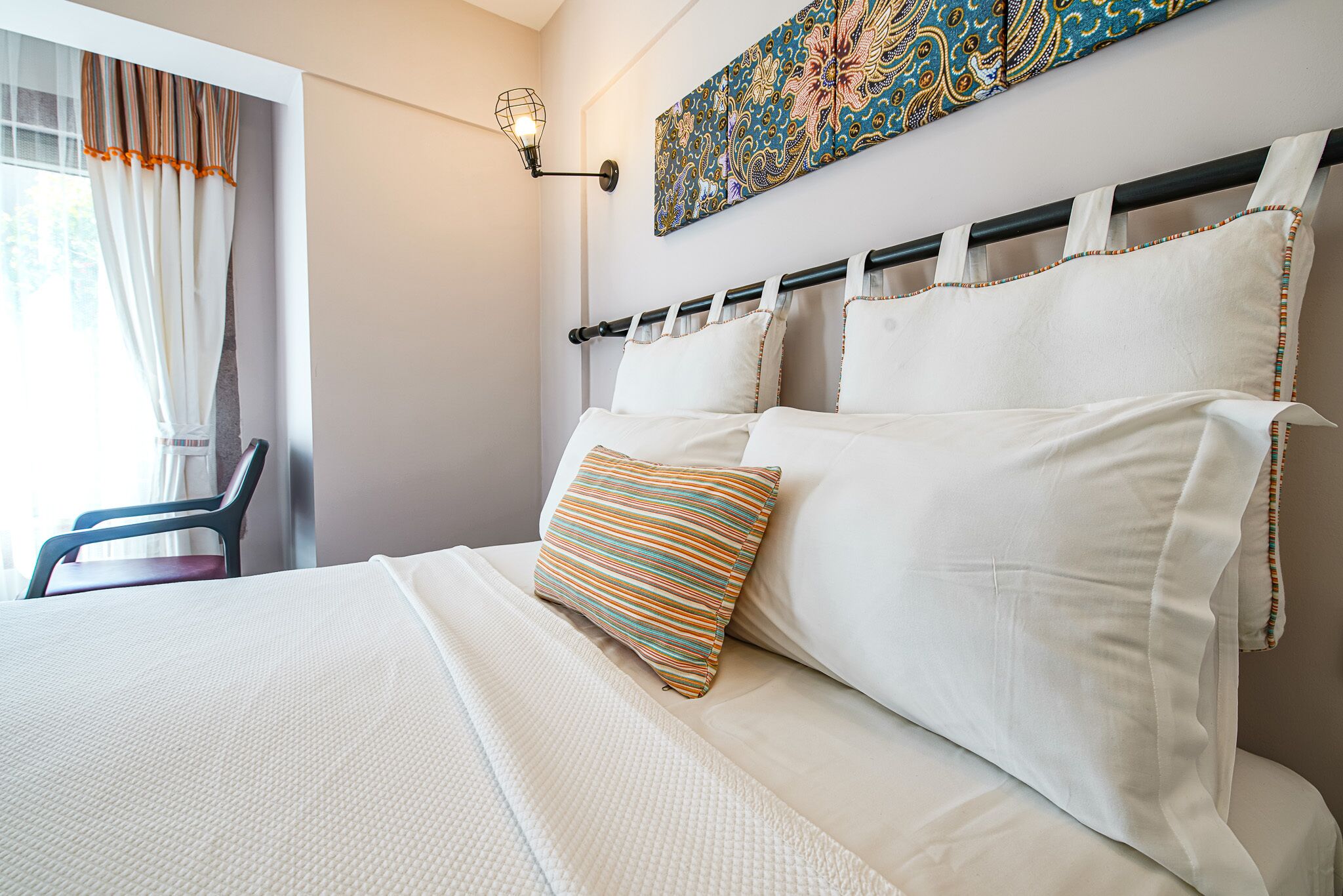 Una cama bien hecha con ropa blanca y un cojín a rayas de colores, una lámpara de pared y un adorno decorativo en la pared.
