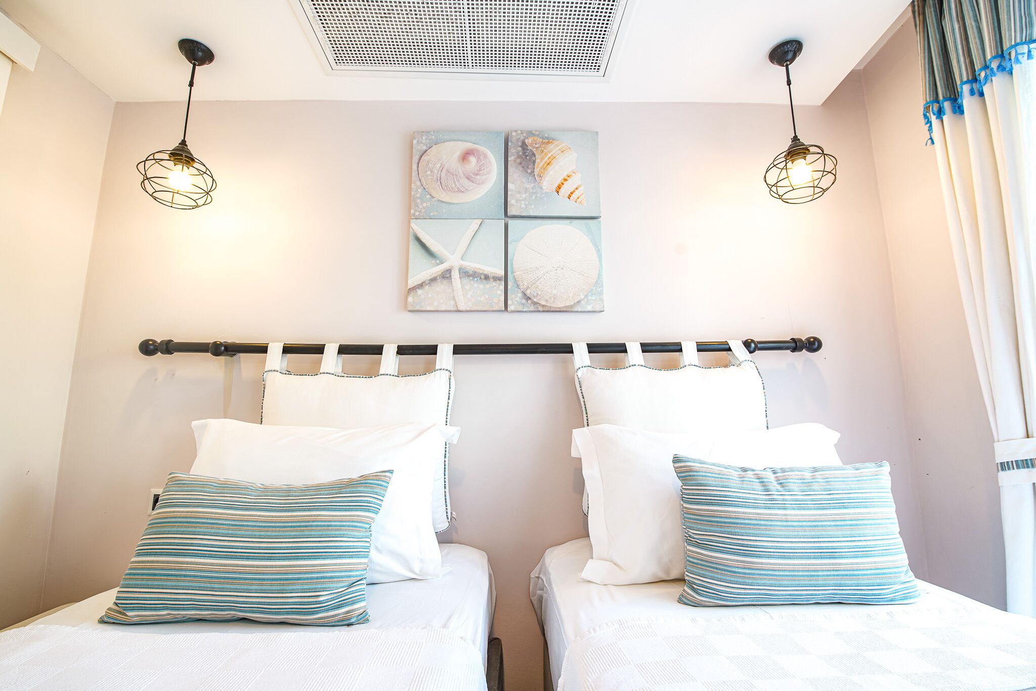 A hotel room with two beds, each with white bedding and striped pillows, a ceiling light fixture, and a wall-mounted artwork depicting seashells and a starfish.