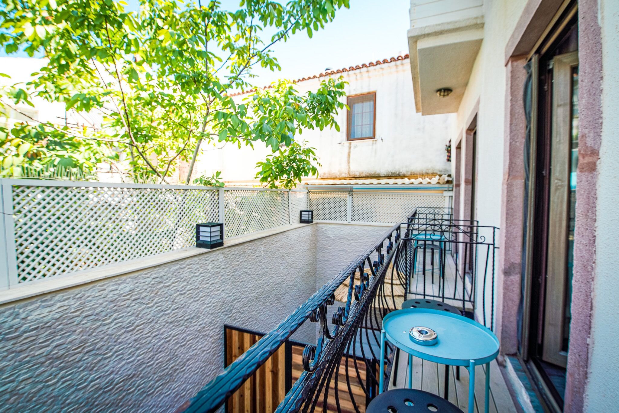 A balcony with a blue table and chairs, a white fence, and a building with a red-tiled roof in the background.