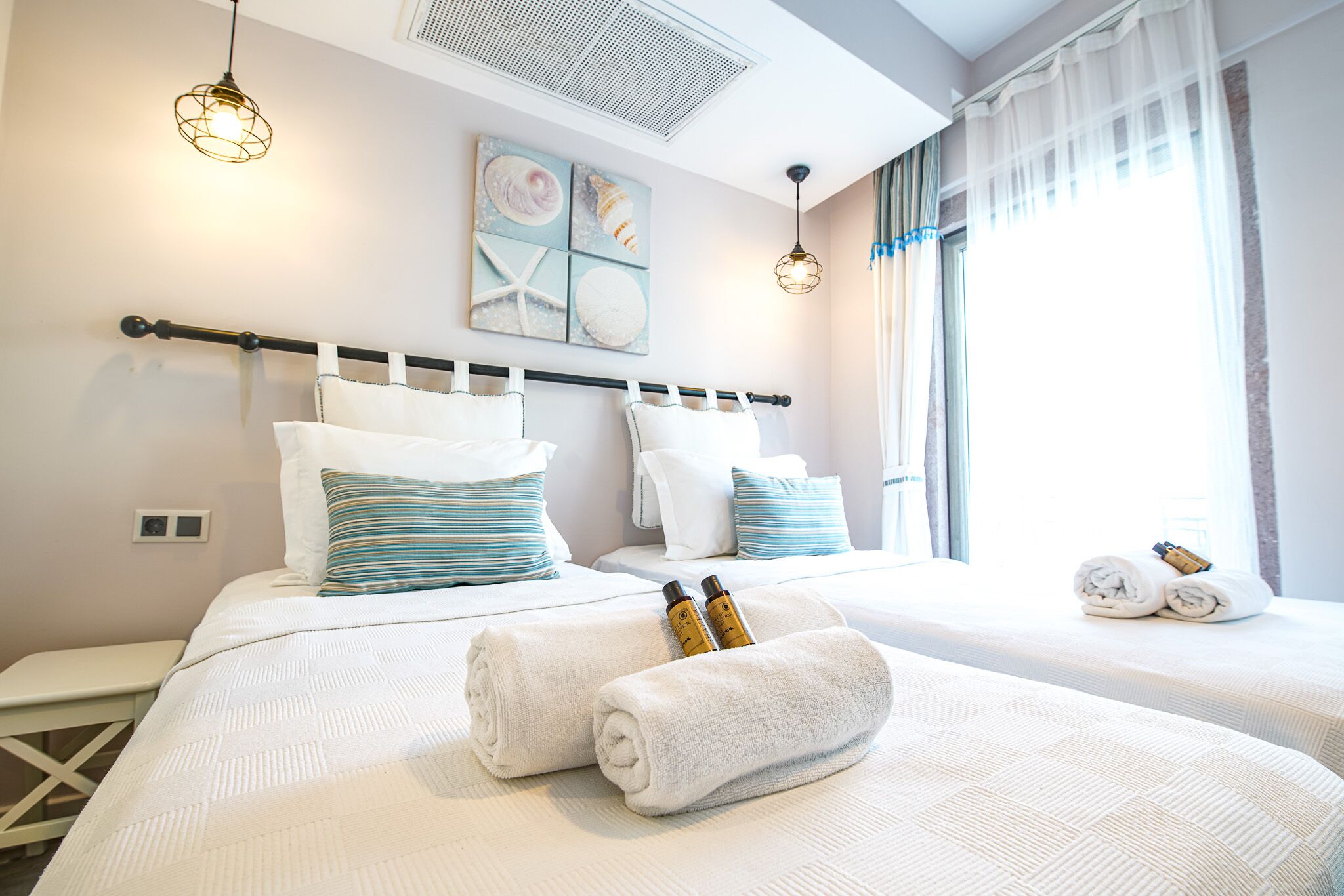 Une chambre d’hôtel avec deux lits, du linge de lit blanc, une tête de lit dotée d’une barre pour les serviettes, un tableau décoratif au mur et une fenêtre avec des rideaux.