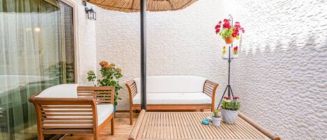 Conjunto de mesa e banco de madeira, com planta pendurada e guarda-sol trançado, em um ambiente com paredes brancas e textura.
